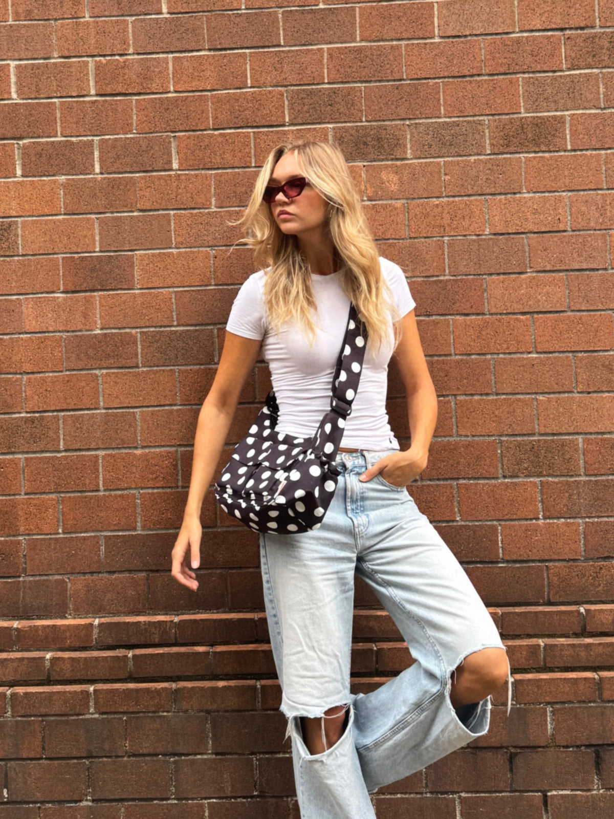 A woman posing against a red-brown brick wall wearing a white t-shirt, light-wash ripped jeans, white sneakers, and sunglasses, while carrying a black and white polka-dot shoulder bag.