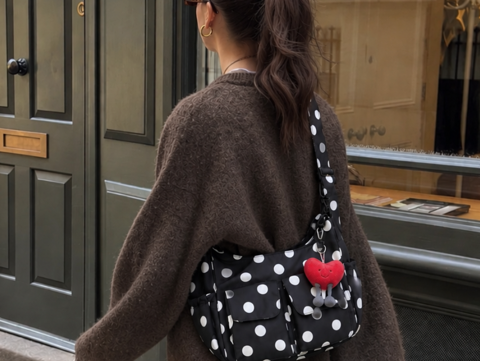 A woman walking on a city street wearing a brown knit sweater, leopard-print wide-leg pants, yellow sneakers, and a black polka-dot crossbody bag with white dots and a red heart accent.